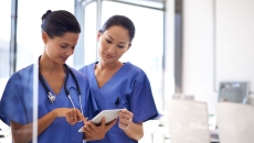   Two healthcare providers in scrubs standing side-by-side looking at a tablet 