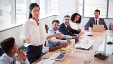 Person standing up at a table in a meeting room with other people sitting down and looking at them
