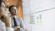 Two people standing up and looking at a large screen with graphs on it