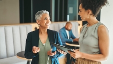 Two individuals walking in an office while another one is sitting in the background