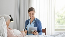Person lying in bed with a healthcare provider sitting beside them in a chair holding a tablet
