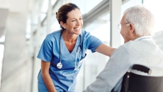 A nurse smiling and standing in front of a window leaning over a patient in a wheelchair