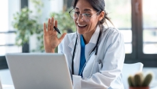 A healthcare provider in a lab coat with a stethoscope around their neck sitting at a desk looking at a computer and waving