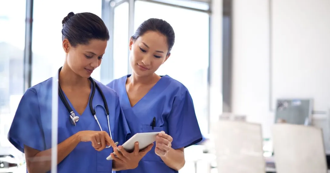   Two healthcare providers in scrubs standing side-by-side looking at a tablet 