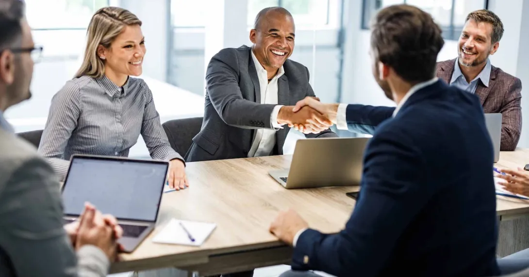 Five businesspersons sitting around a table with two of them shaking hands