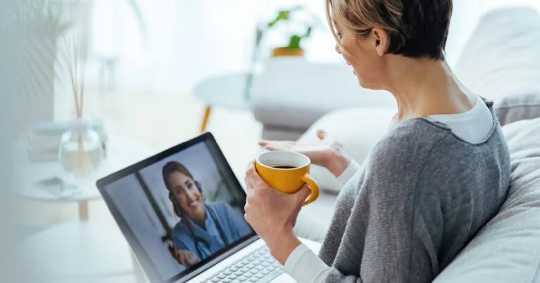 Person sitting on their couch holding a cup of coffee while speaking to a virtual healthcare provider on their computer