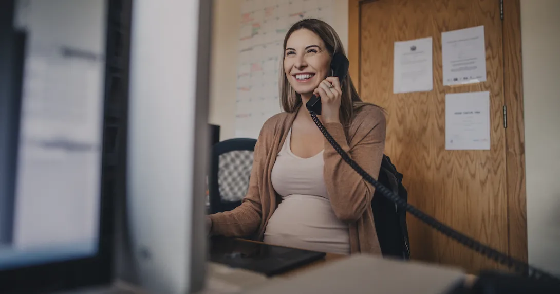 Pregnant person sitting at a desk talking on the phone while looking at a computer