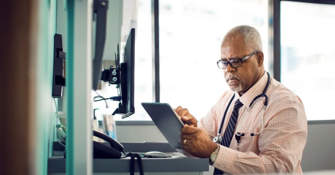 "Healthcare professional sitting at a desk with a window behind them while wearing a stethoscope around their neck and looking at a tablet"