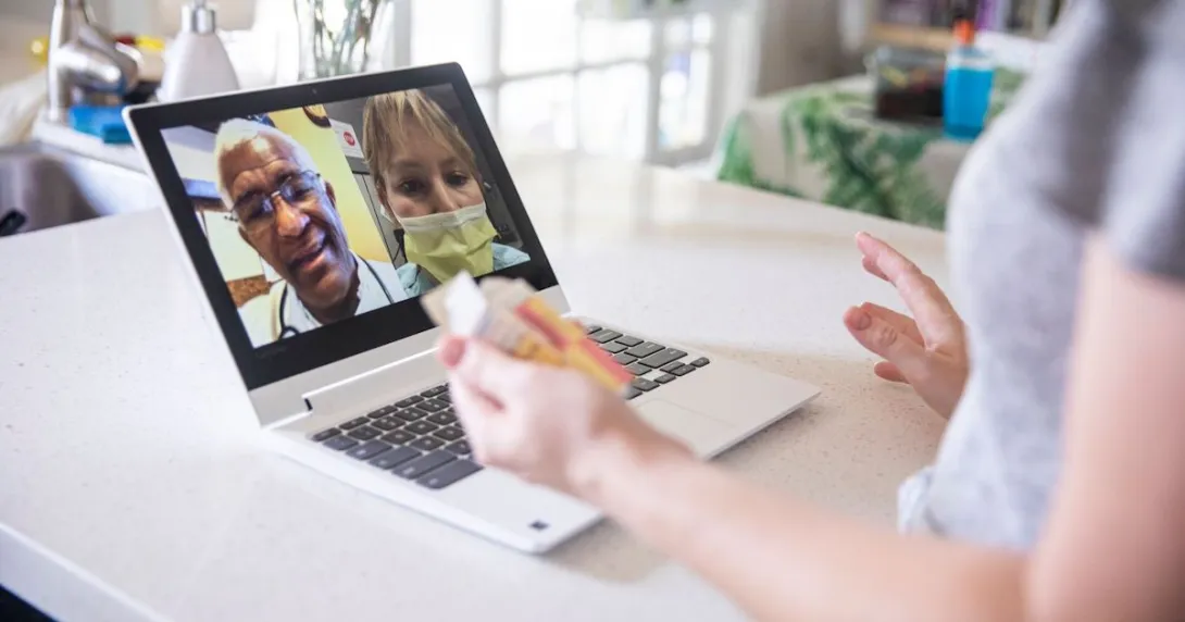 A close up of a woman holding a prescription while talking to two providers on her laptop
