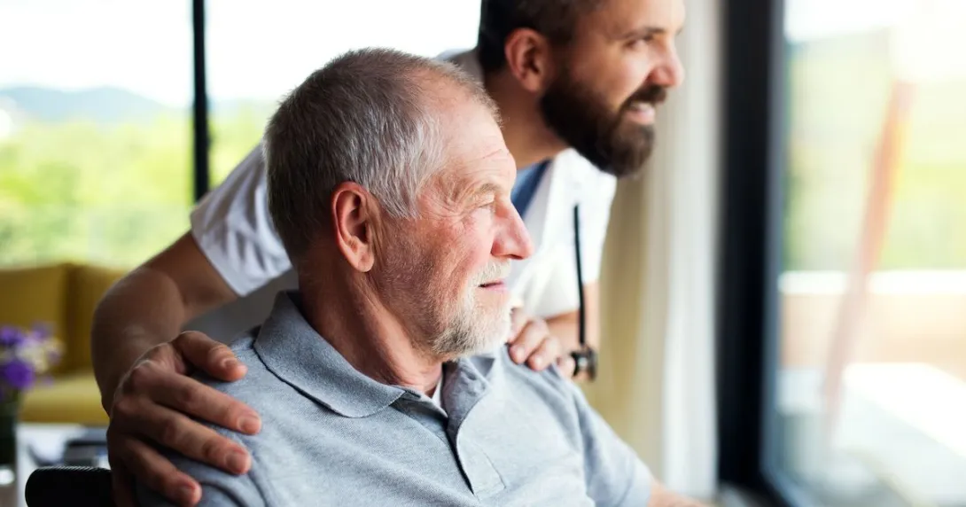 Person sitting in a chair looking out a window with a healthcare provider next to them with their hands on the person's shoulders