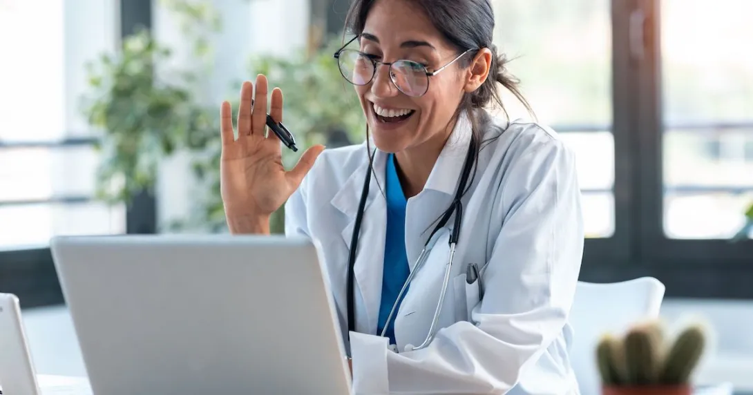 Healthcare provider sitting at a desk and looking at a computer while waving to the screen