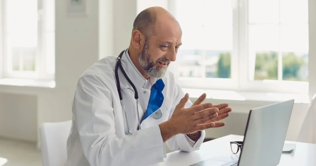 Healthcare provider wearing a lab coat and stethoscope while sitting at a desk and talking to someone on a computer
