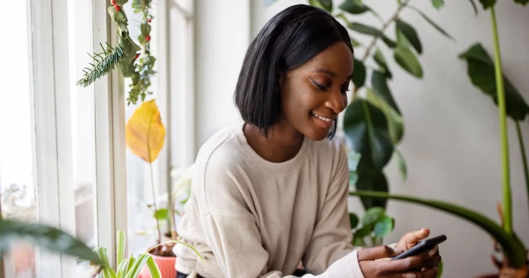A person using a smartphone sitting by a window and surrounded by plants